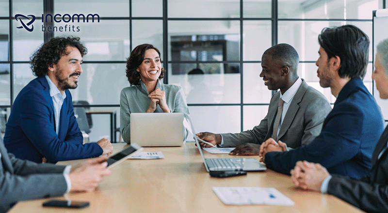 Diverse business team in a meeting around a wooden table with laptops, smiling; Incomm Benefits logo in the top-left.