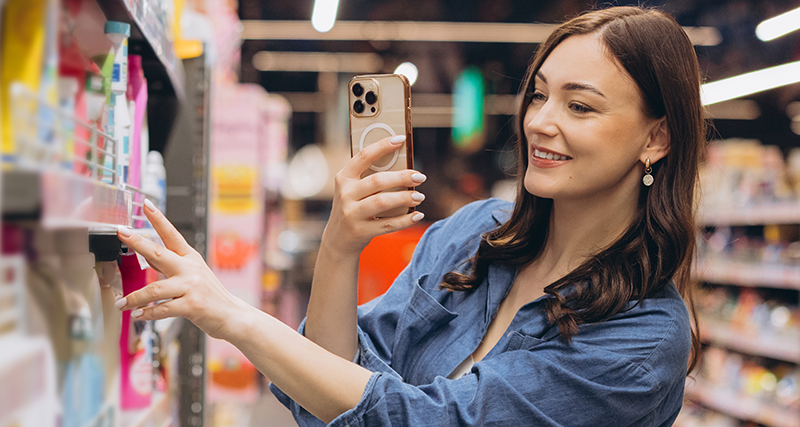 Woman in a blue shirt shops in a store aisle, taking a selfie with her phone as she reaches for a product on the shelf.