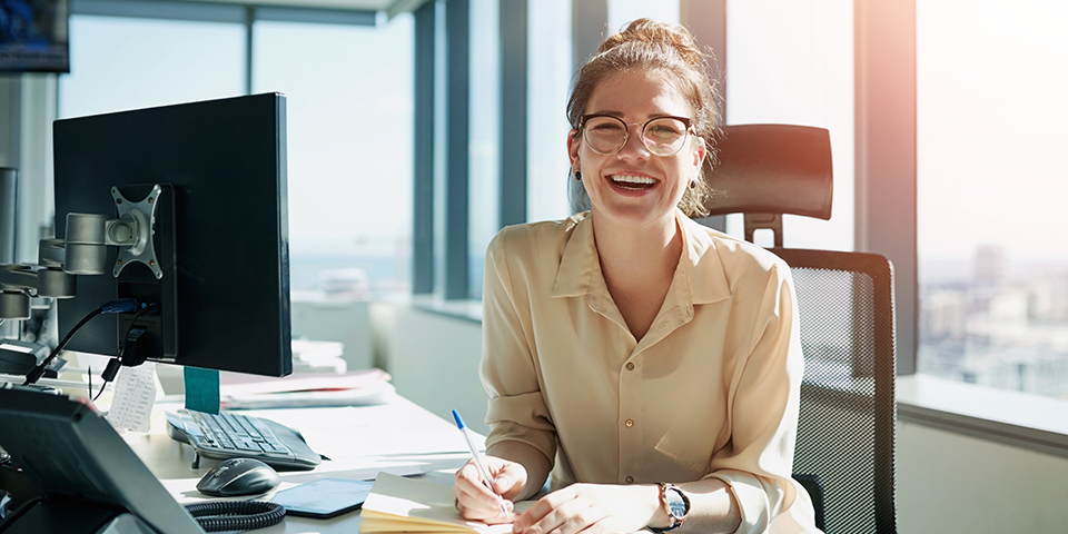 Smiling woman in a beige blouse writes at a desk in a bright office with a computer monitor and city view outside the window.