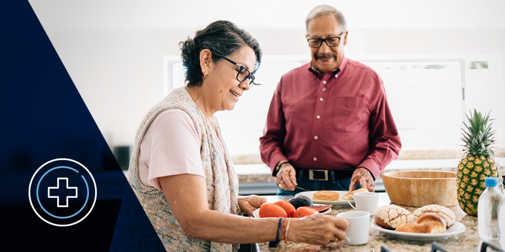 Senior couple cooking together in a bright kitchen; woman chops tomatoes while man stands nearby with bread and fruit on the counter, smiling as they prepare a meal.